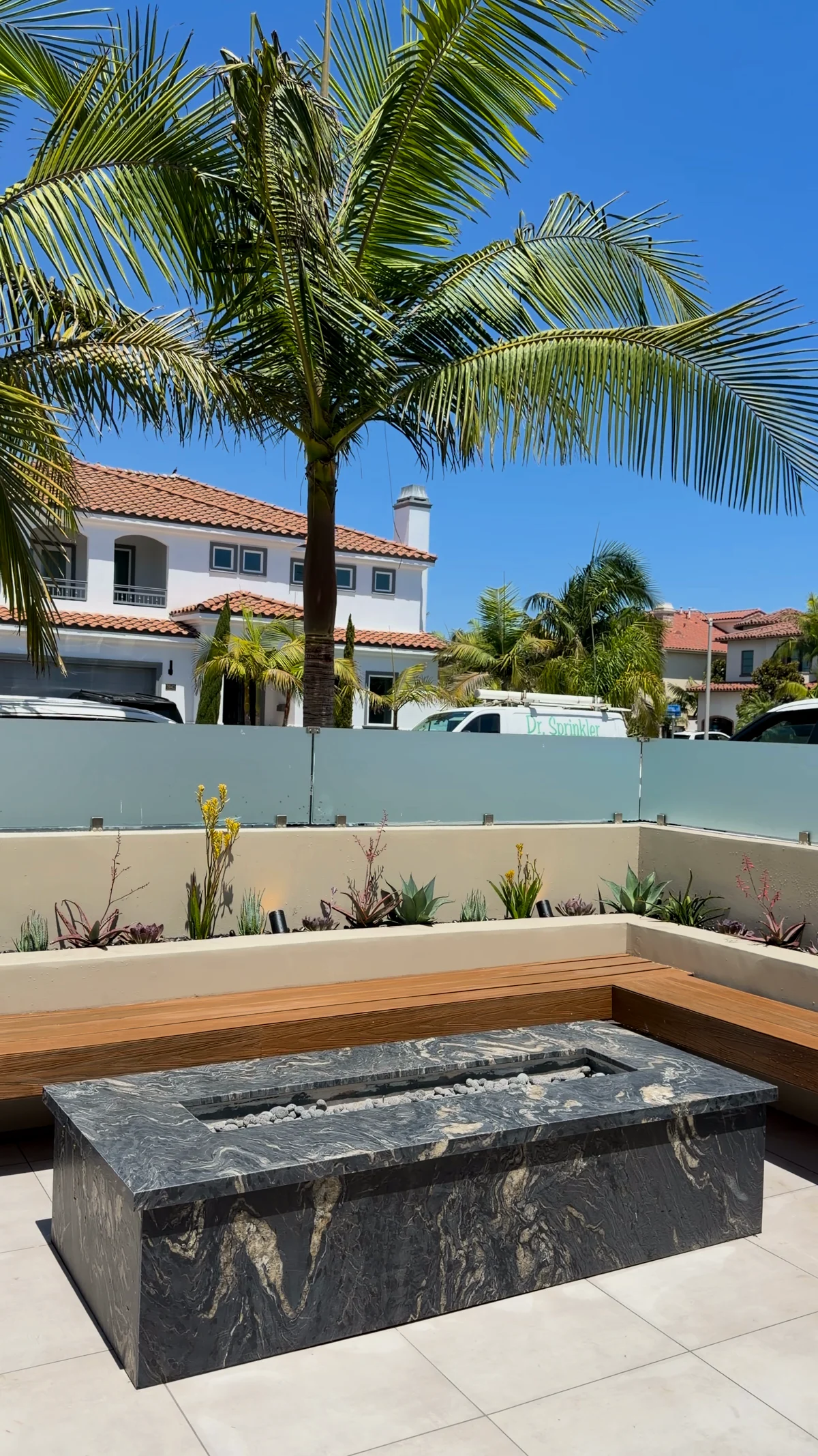 A cantilevered teak bench wrapping a courtyard with a solid stone fire table on large format pale porcelain paving