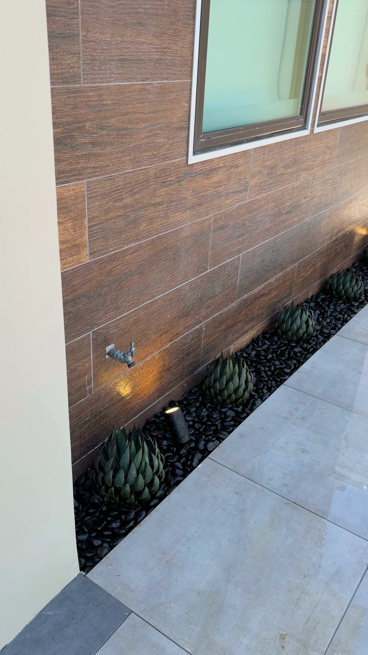 A narrow side yard with a wood look porcelain plank wall, agaves set in black pebble, and a low uplight washing the wall