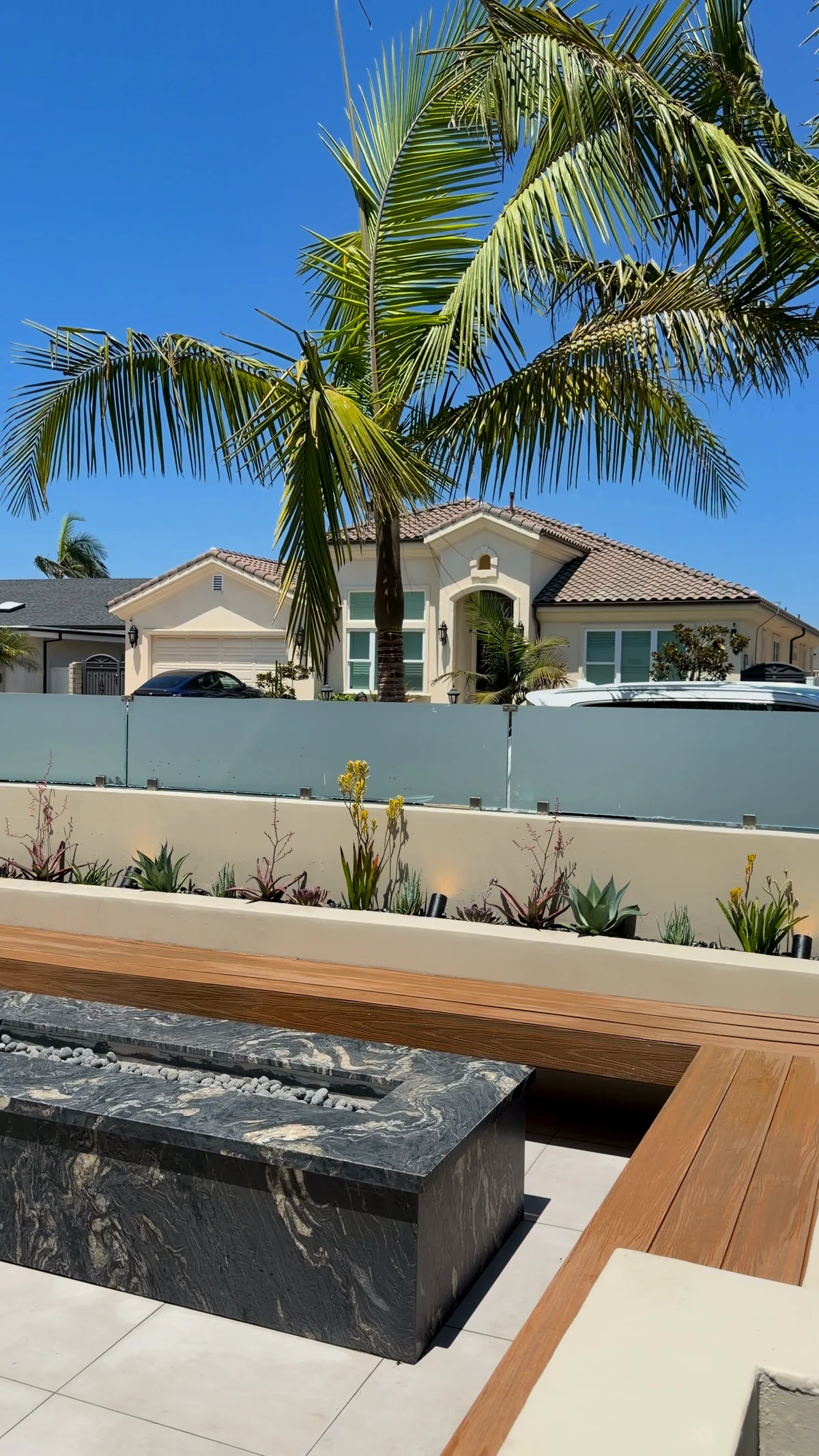 The finished coastal courtyard seen wide, with glass wind panels, planted beds, a wrapping bench and a stone fire table under queen palms